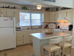 A kitchen with white cabinets, a refrigerator, a microwave, a toaster, and a sink. There are two chairs at the island and a window with blinds. - School Bored --Affordable Cottage just 2 blocks to the ocean (Kill Devil Hills)