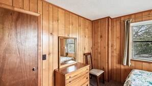 Ein Schlafzimmer mit Holzwandverkleidung, einer Kommode mit Schubladen, einem Sessel, einem Bett mit gemustertem Bettbezug und einem Fenster mit Jalousien.