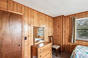 A bedroom with wooden panel walls, a dresser with drawers, a chair, a bed with a patterned bedspread, and a window with blinds. - Owl's Nest -- Vacay Your Way with Flexible Arrival and Stay Dates (Kill Devil Hills)