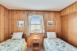 Une pièce aux murs en bois, avec deux lits dotés de draps à motifs floraux, une table de chevet avec une lampe et une fenêtre avec des rideaux.