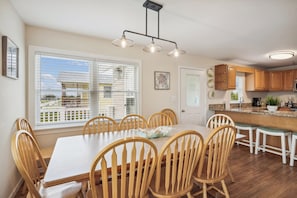 Un coin repas avec une table blanche et des chaises en bois, une cuisine avec des meubles en bois et une fenêtre équipée de stores.