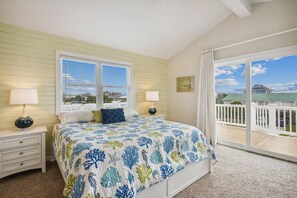 A bedroom with a large bed, two bedside tables, a window with blinds, a view of the outside, and a balcony with a railing. - Gone to Carolina -- Recently Remodeled Home with Pool, Hot Tub, & Putting Green (Kitty Hawk)