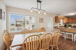 A dining area with a table and chairs, a kitchen with wooden cabinets, and a window with blinds. - Gone to Carolina -- Recently Remodeled Home with Pool, Hot Tub, & Putting Green (Kitty Hawk)