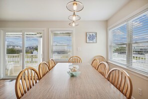 Une salle à manger avec une longue table en bois, six chaises en bois, un lustre et de grandes fenêtres équipées de stores.