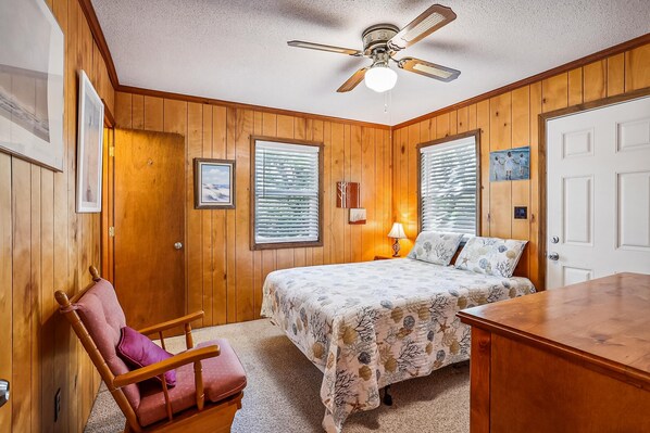 A bedroom with wooden paneling, a bed with a floral bedspread, a chair, a desk, and a ceiling fan.