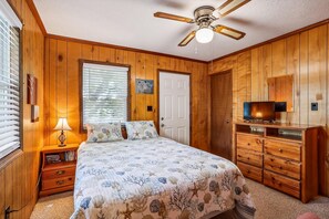 A bedroom with wooden paneling, a bed with floral bedding, a ceiling fan, a television on a wooden cabinet, and a window with blinds.