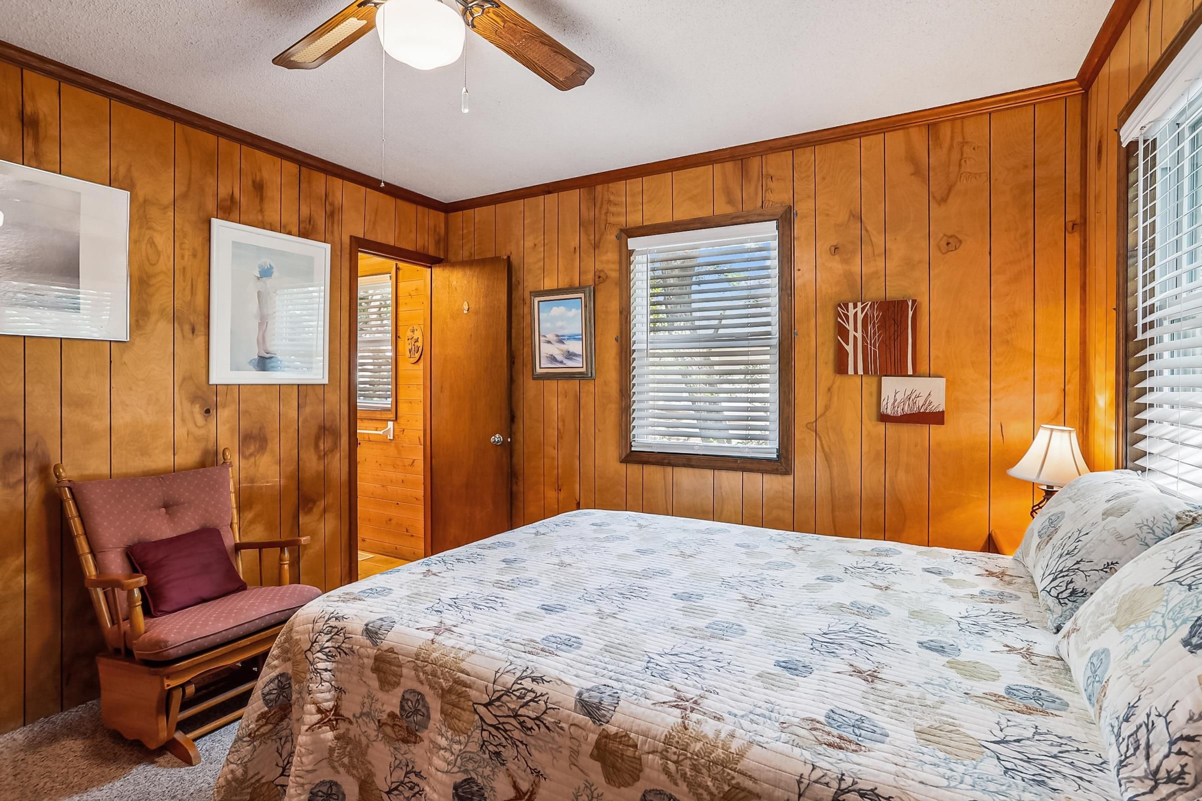 A bedroom with wooden walls, a bed with a floral bedspread, a chair, a ceiling fan, and a window with blinds.