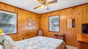Een slaapkamer met houten lambrisering, een bed met een bloemenprint sprei, een plafondventilator en een televisie op een houten dressoir.