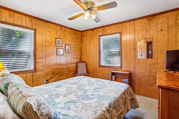 Une chambre avec des lambris en bois, un lit recouvert d’une couvre-lit à motifs floraux, un ventilateur de plafond et un téléviseur posé sur un meuble en bois.