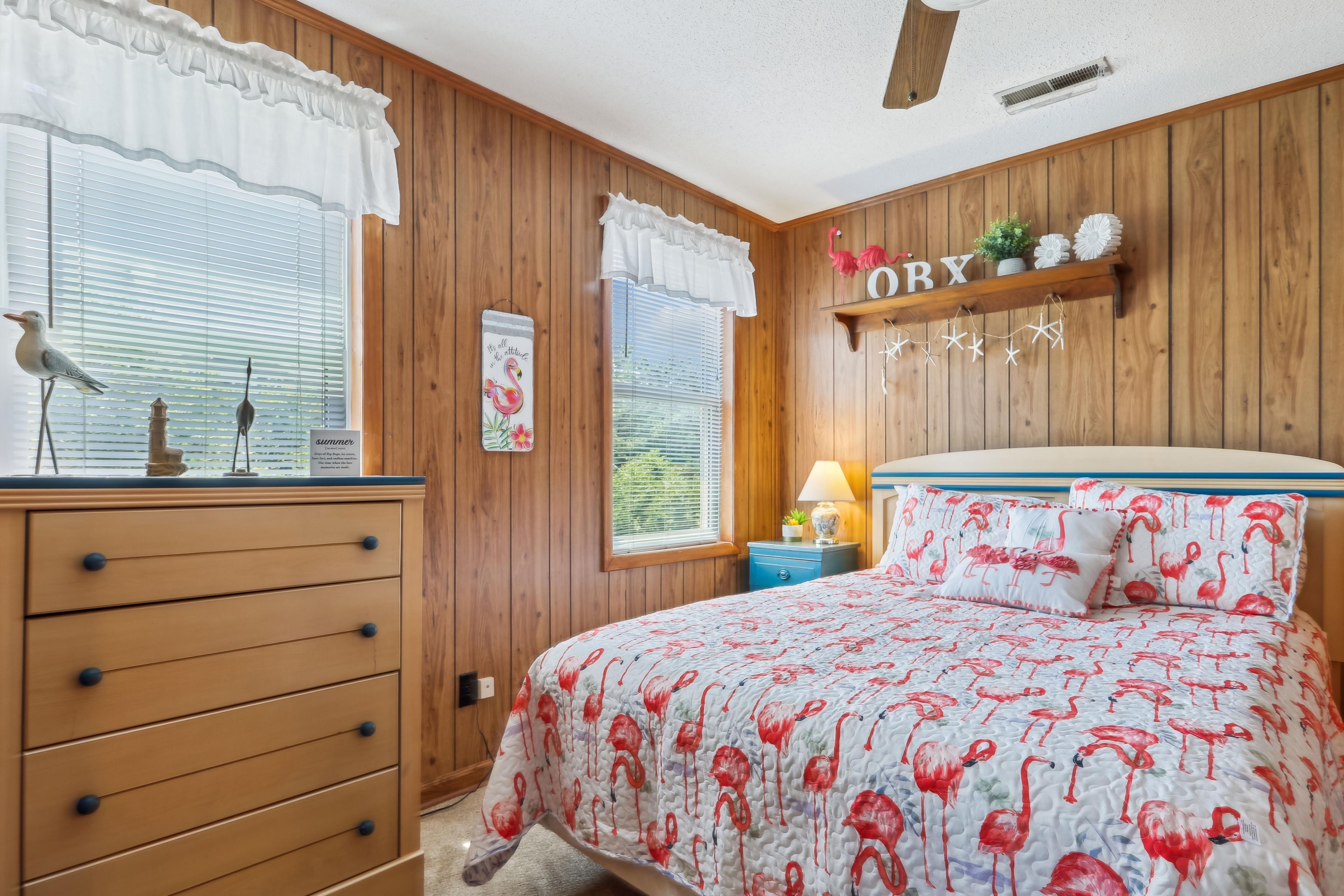A bedroom with a wooden paneled wall, a bed with a flamingo-themed bedspread, a dresser with drawers, and a window with a bird figurine.