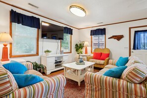 Una sala de estar con sofás a rayas, una mesa de centro blanca, una silla de mimbre con cojines rojos y un televisor montado en la pared.