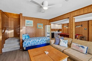 A room with a bed, a sofa, a coffee table, a ceiling fan, wooden walls, a staircase, and a kitchen area.