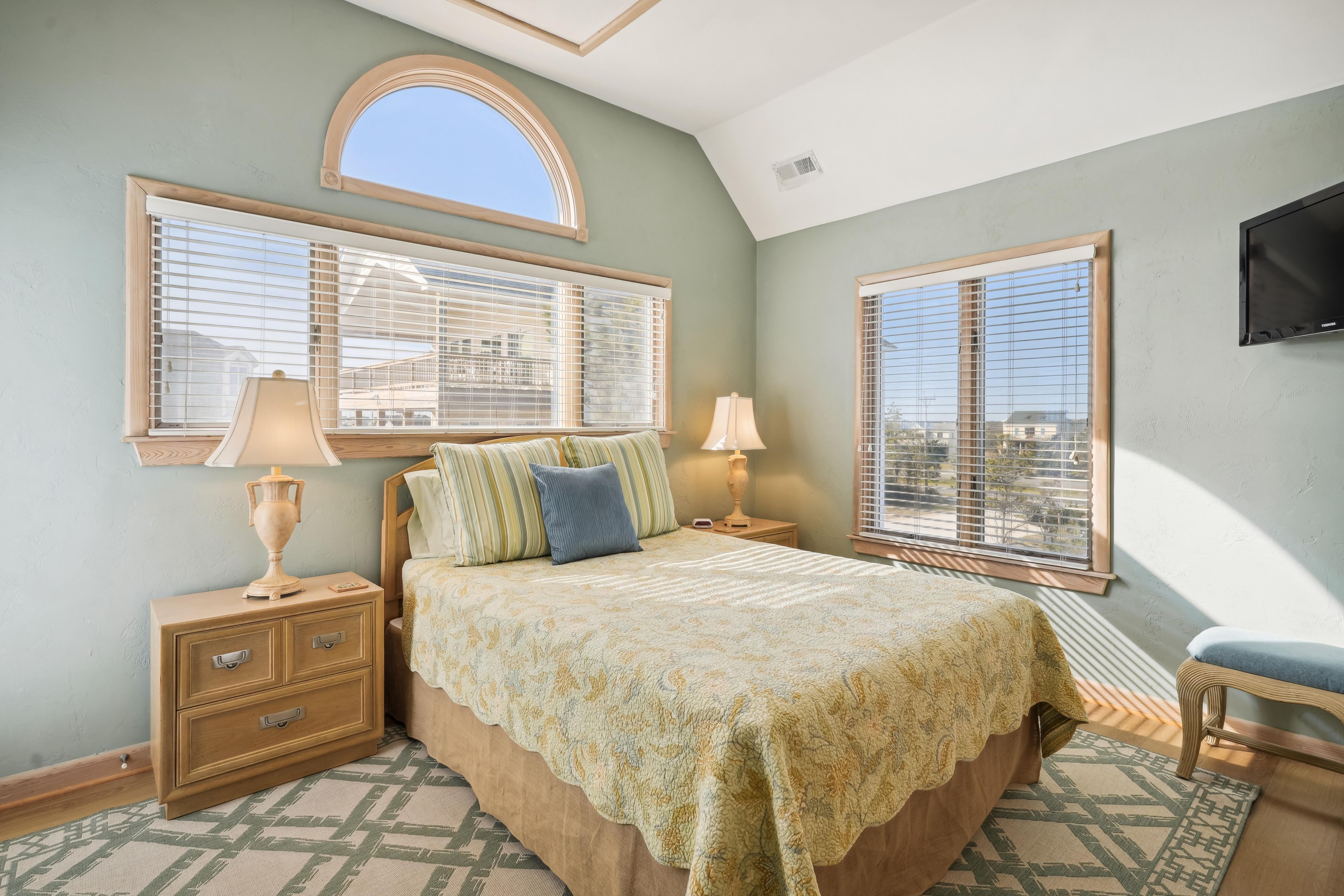 A bedroom with a bed, two bedside tables, a nightstand, a TV mounted on the wall, and large windows with blinds.