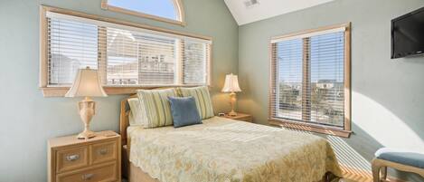 A bedroom with a bed, two bedside tables, a nightstand, a TV mounted on the wall, and large windows with blinds.