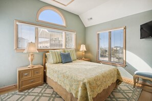 A bedroom with a bed, two bedside tables, a nightstand, a TV mounted on the wall, and large windows with blinds.