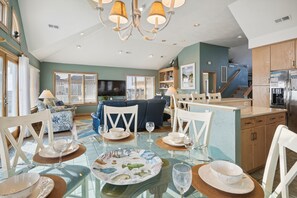 Een ruime woonkamer met een eettafel voor vier personen, een blauwe bank en een keuken met een koelkast en fornuis.