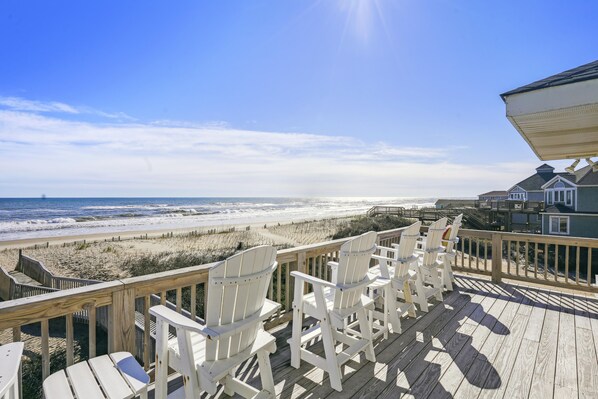 A wooden deck with white chairs overlooking a beach and ocean.