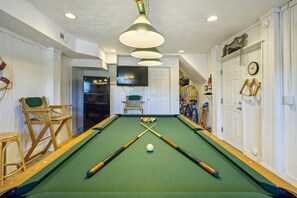 A pool table with two cues and a white ball, a television mounted above it, and a chair with a cushion.