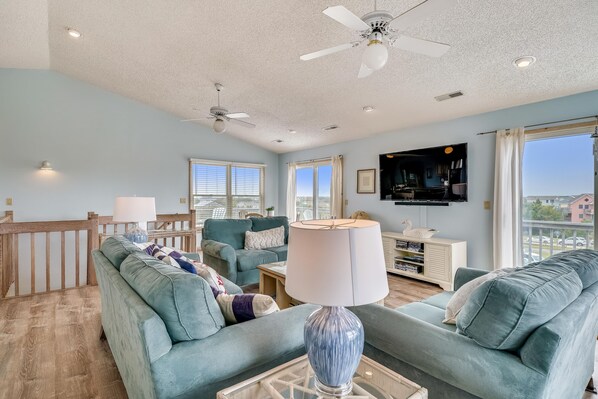 A living room with a ceiling fan, a TV mounted on the wall, and a large window with curtains. - Sea Duck Sun -- Dog Friendly 5BR in Downtown Duck (Duck)