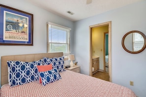 A bedroom with a bed, a nightstand, a lamp, a framed picture, a window with blinds, and a mirror.
