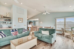 A living room with a green sofa, a wooden coffee table, and a dining area with wicker chairs. - Sea Duck Sun -- Dog Friendly 5BR in Downtown Duck (Duck)
