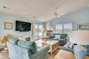 A living room with a flat-screen TV, a ceiling fan, and a large window with blinds. - Sea Duck Sun -- Dog Friendly 5BR in Downtown Duck (Duck)