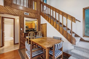 Un escalier en bois avec un palier recouvert de moquette, un coin repas avec une table en bois et des chaises à rayures, et une cuisine visible à travers une porte ouverte.