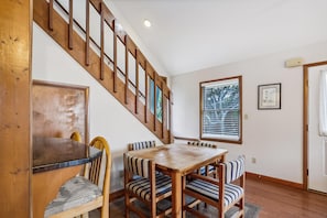 A dining area with a wooden table and chairs, a staircase, and a window with blinds. - Keepers Loft -- Waterfront Duplex with Pier and Gazebo (Duck)