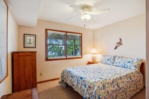 A bedroom with a bed, a nightstand, a lamp, a ceiling fan, a window with blinds, and a wall-mounted bird decoration.