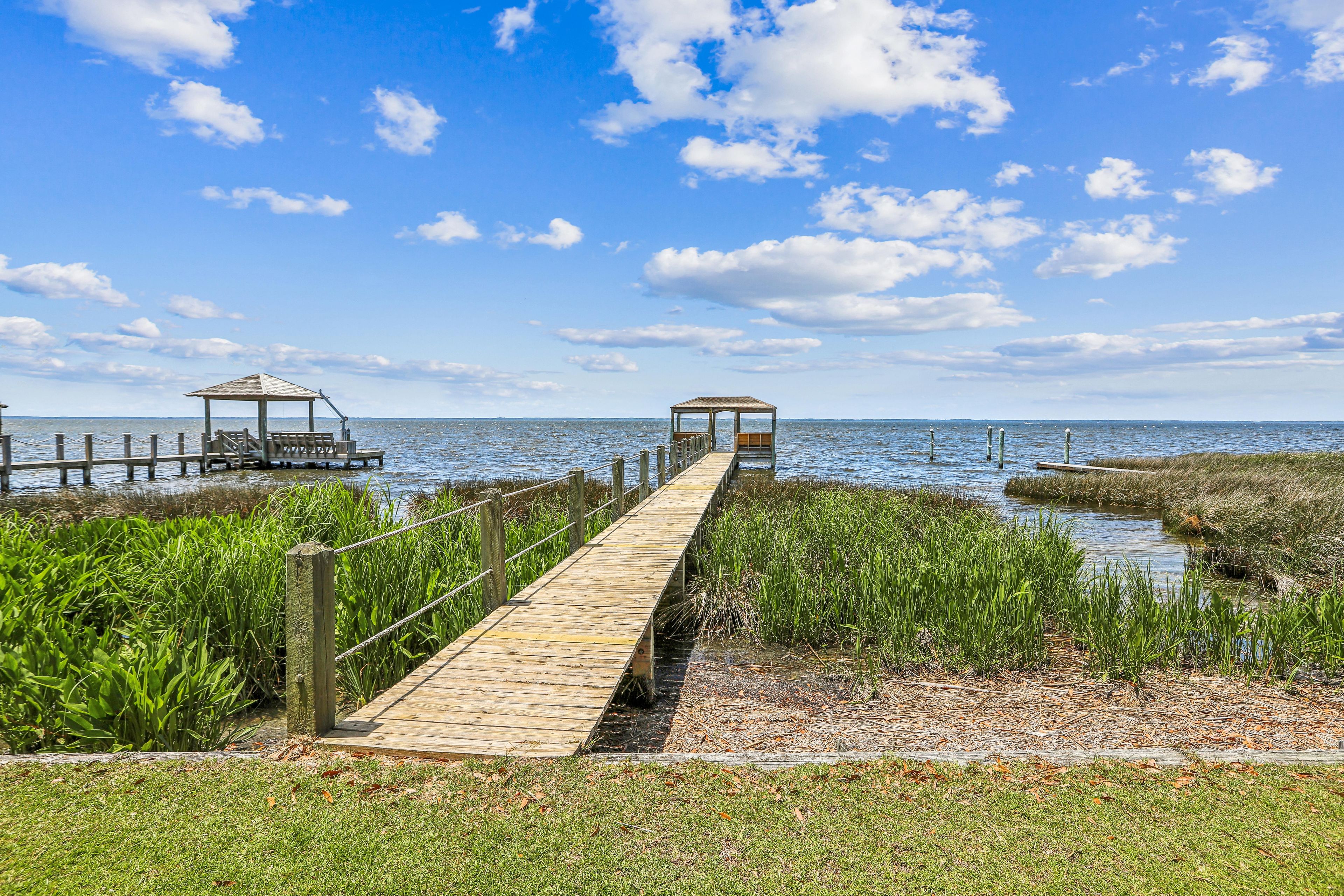 A wooden boardwalk leading to two gazebos over water, surrounded by grassy areas.