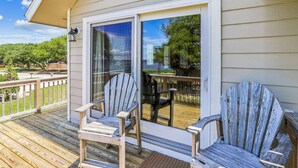 A wooden deck with two Adirondack chairs and a glass door leading to a patio area. - Emerald Duck -- Awesome Sunset and Water Views Soundfront Duplex (Duck)