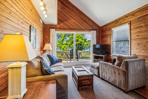 A cozy room with wooden walls, a sofa, a chair, a TV, and a window with a view of trees.