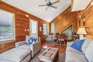 A cozy living room with wooden walls, a ceiling fan, and a dining area with wicker furniture. - Emerald Duck -- Awesome Sunset and Water Views Soundfront Duplex (Duck)