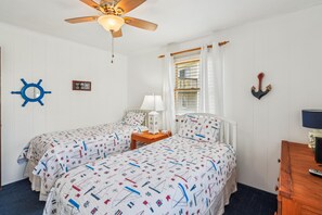 A room with two beds, a ceiling fan, a window with curtains, and nautical-themed bedding.