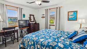 A bedroom with a bed, a desk, a television, a ceiling fan, and a window with curtains.