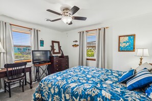 Un dormitorio con cama, escritorio, televisor, ventilador de techo y ventana con cortinas.