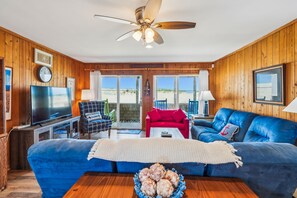 Una sala de estar con un sofá azul, un televisor, un ventilador de techo y vistas a la playa a través de amplios ventanales.