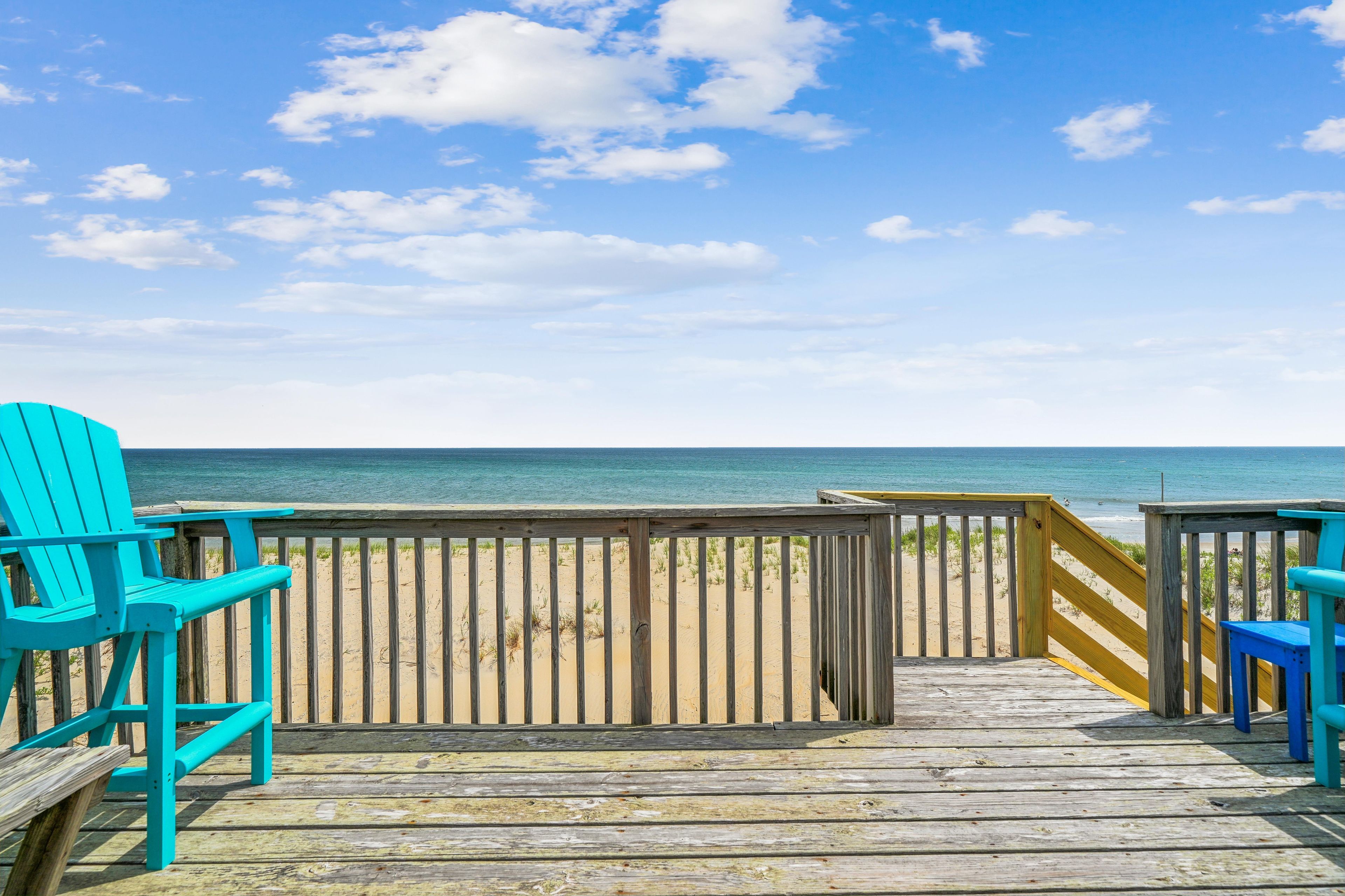 Une promenade en bois mène à une plage où l’on trouve, de part et d’autre, des chaises de couleur turquoise.
