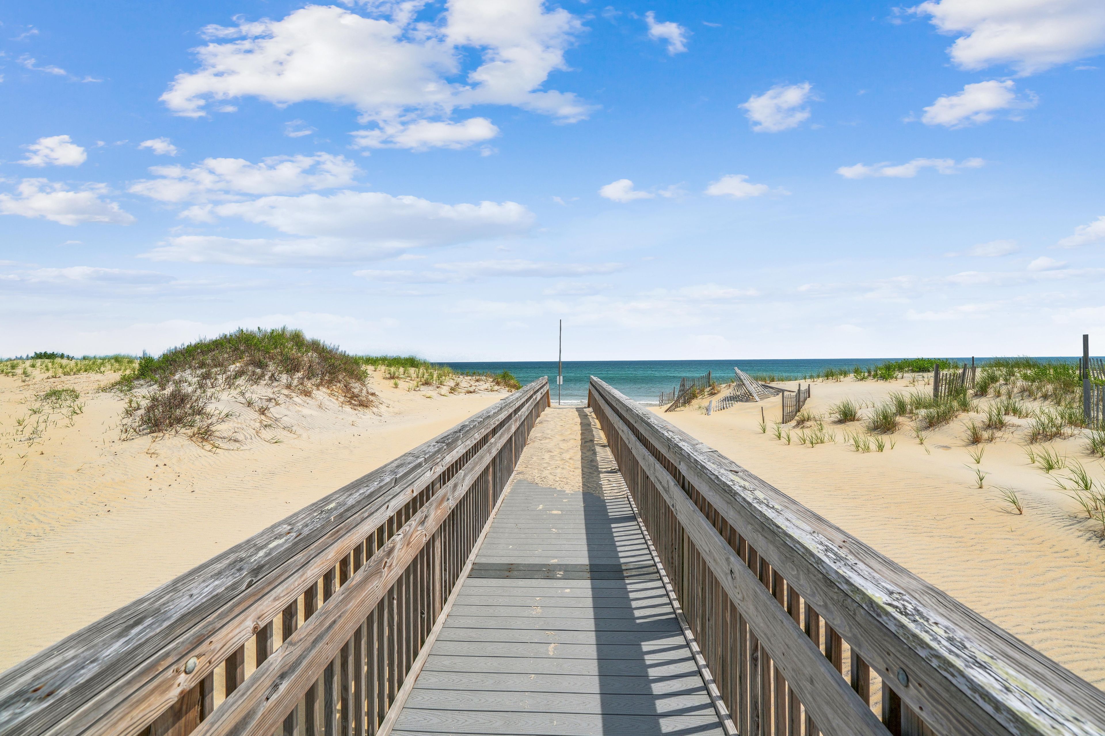 A wooden boardwalk leading to a beach with dunes and a clear sky.