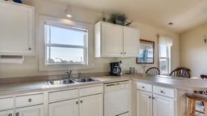 A kitchen with white cabinets, a double sink, a coffee maker, and a dining area with wooden chairs. - Premium Tripp -- Close to Shopping, Restaurants and Activities (Kill Devil Hills)
