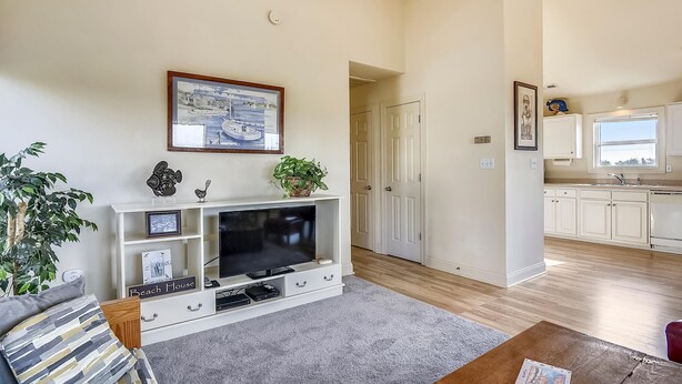 A living room with a flat-screen TV, a coffee table, and a sofa. There is a hallway leading to a kitchen with white cabinets and a window. - Premium Tripp -- Close to Shopping, Restaurants and Activities (Kill Devil Hills)