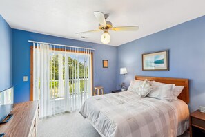 Un dormitorio con cama, mesita de noche, lámpara, ventilador de techo y vistas a través de una ventana con persianas.