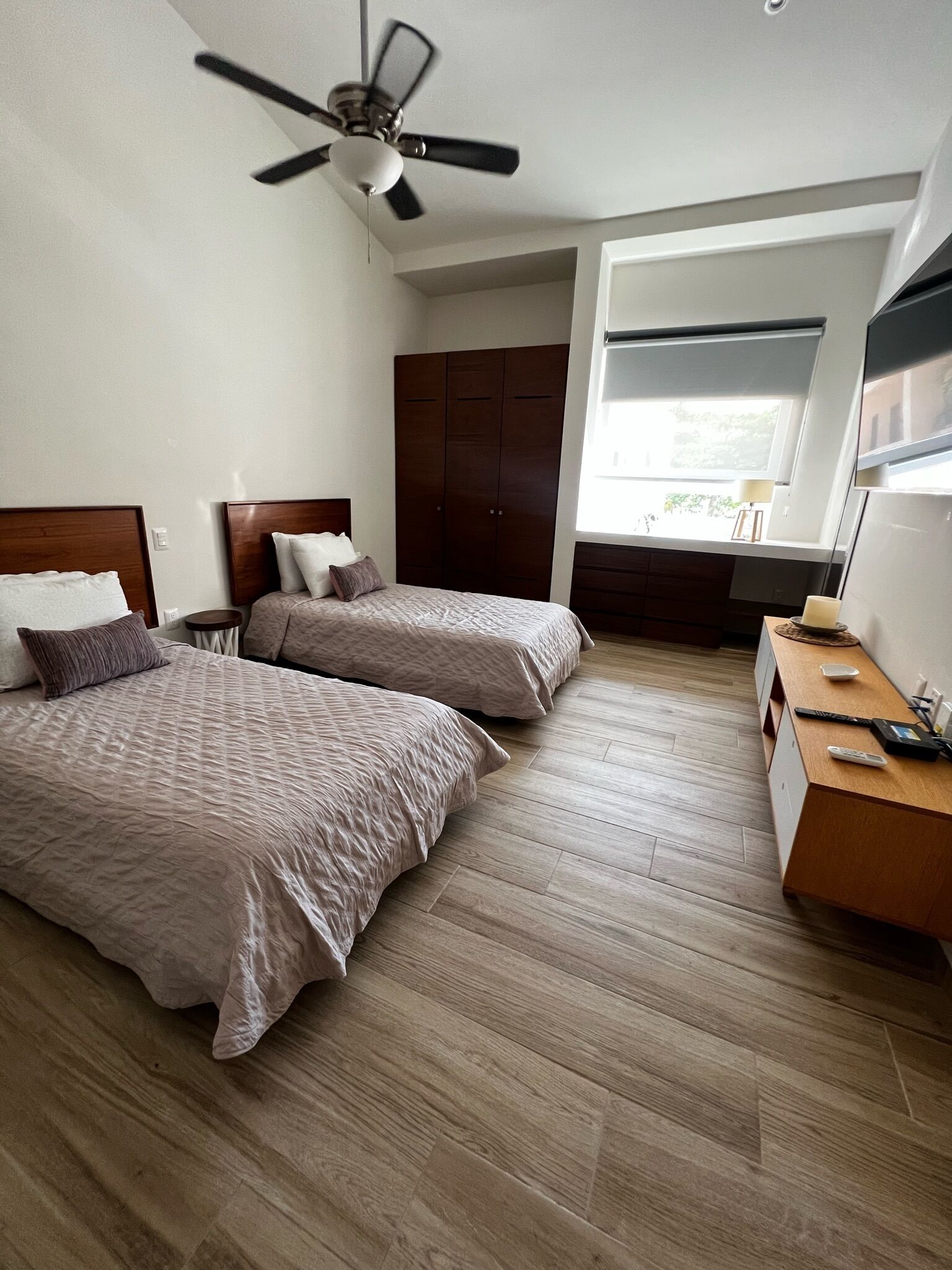 A hotel room with two beds, a wooden floor, a ceiling fan, a wardrobe, a desk with a lamp, and a window with a view.