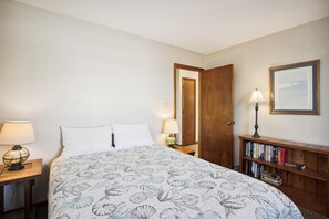 A bedroom with a bed, two bedside lamps, a wooden bookshelf with books, and a door leading to another room.