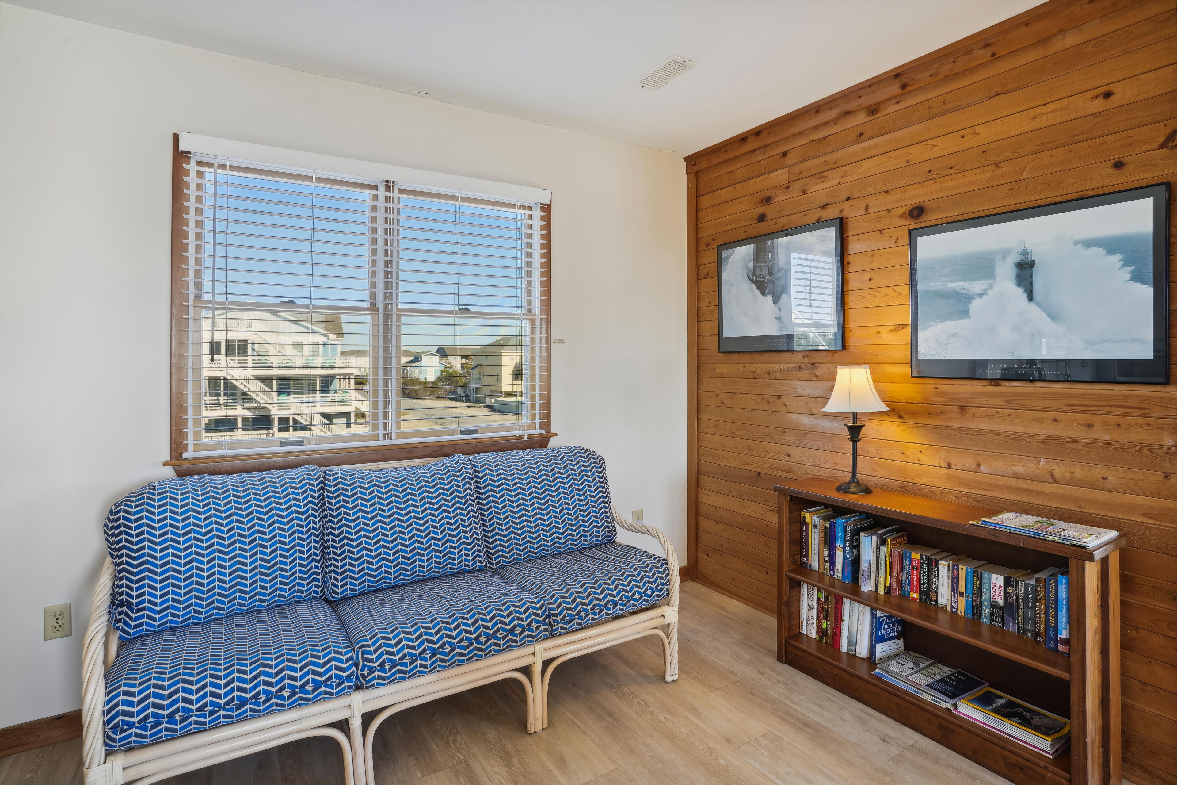 A room with a wooden wall, a window with blinds, a patterned sofa, a bookshelf, and a lamp.