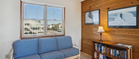 A room with a wooden wall, a window with blinds, a patterned sofa, a bookshelf, and a lamp.