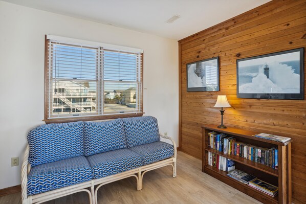 Una habitación con pared de madera, ventana con persianas, sofá estampado, una estantería y una lámpara.