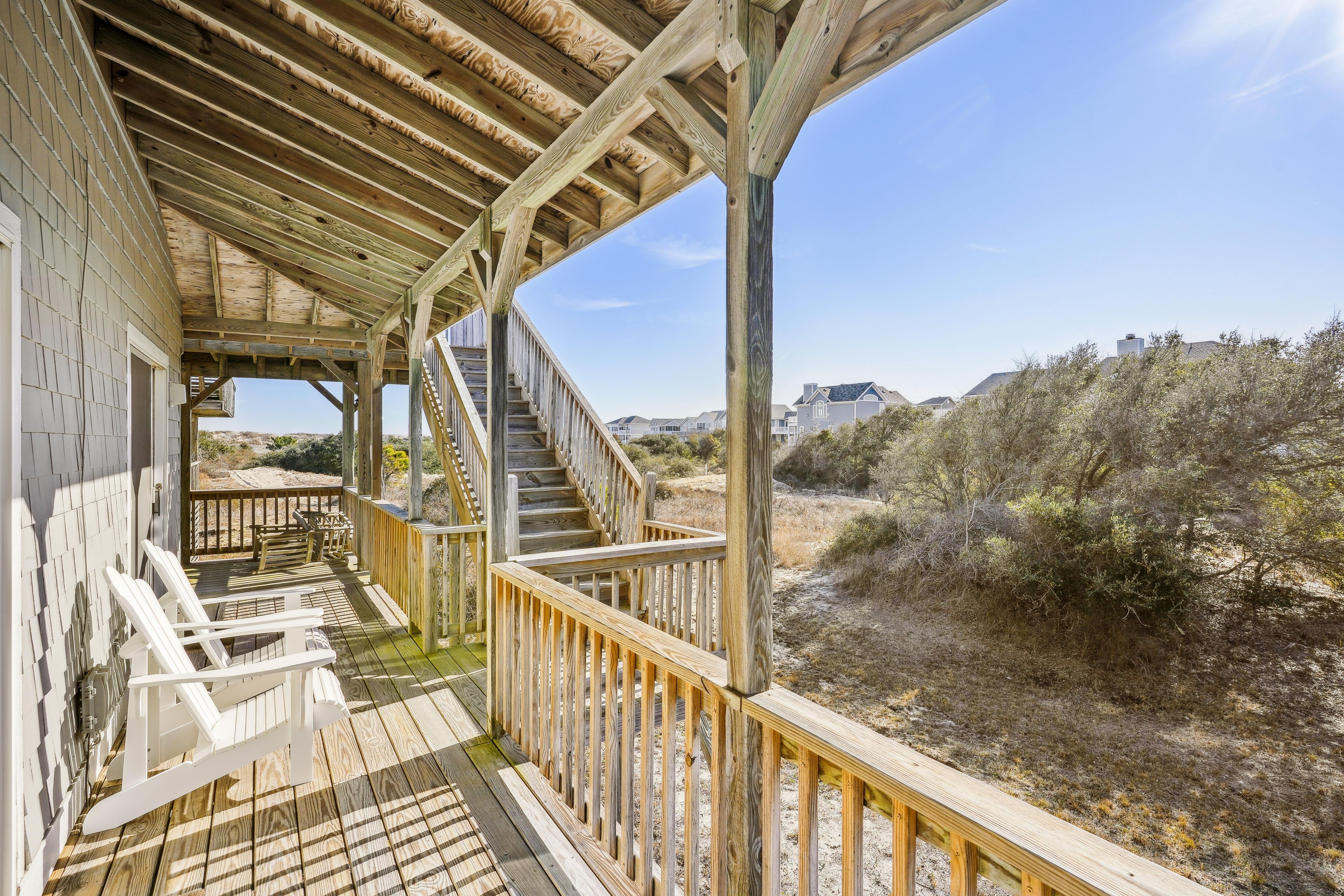 A wooden deck with a staircase leading to a covered area, surrounded by dry vegetation and distant buildings.