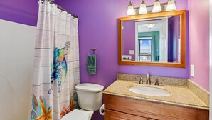 Une salle de bain avec un mur violet, un meuble-lavabo en bois avec un évier, un miroir et un rideau de douche aux motifs colorés.