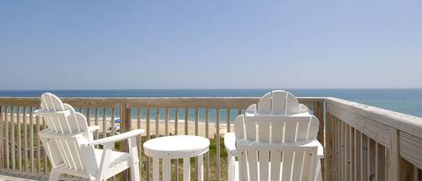 A wooden deck with white Adirondack chairs and a small round table, overlooking a beach and ocean.